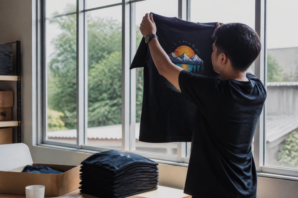 A person holding a custom-printed t-shirt up to a window light, inspecting the print quality closely in a bright room