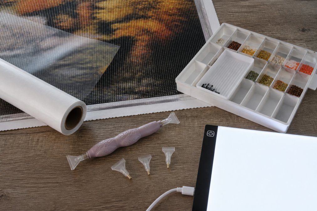 Flat lay of premium diamond painting tools including ergonomic dual-head drill pen with multi-placer attachments, light pad illuminated with adjustable brightness, organized drill tray with compartments, and release paper roll, arranged on a workspace with a canvas in progress showing partial completion
