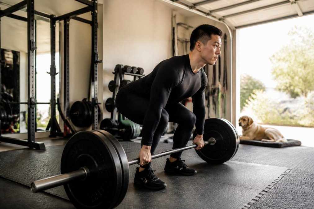 A lifter in fitted athletic compression tights and a snug training top setting up for a deadlift in a home garage gym, dog resting on a mat nearby, warm afternoon light through an open door