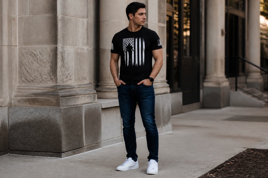 Man in a black patriotic graphic t-shirt, dark jeans, and white sneakers posing in an urban streetwear setting.