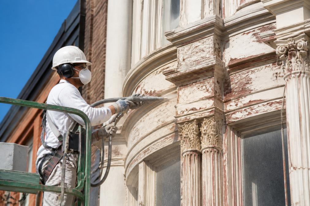 Soca Services professional performing careful paint removal on heritage building facade with specialized equipment, demonstrating precision surface treatment in bright daylight