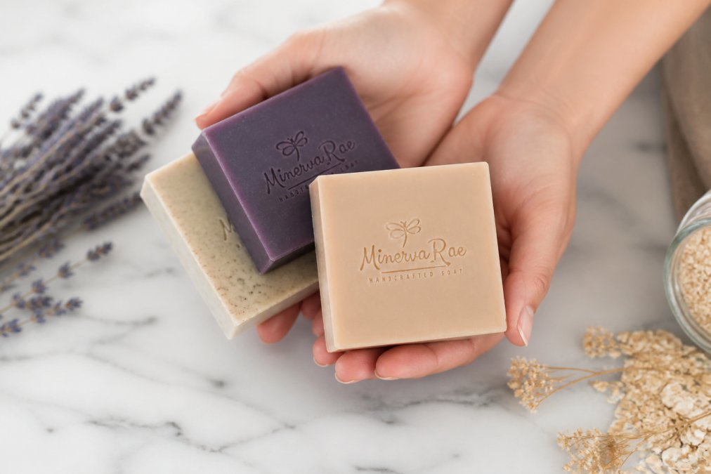 Close-up of hands gently holding three handcrafted soap bars in natural lighting, showing creamy white unscented bar, deep purple lavender bar, and oatmeal-speckled bar arranged on a marble surface with dried lavender sprigs and botanical elements nearby