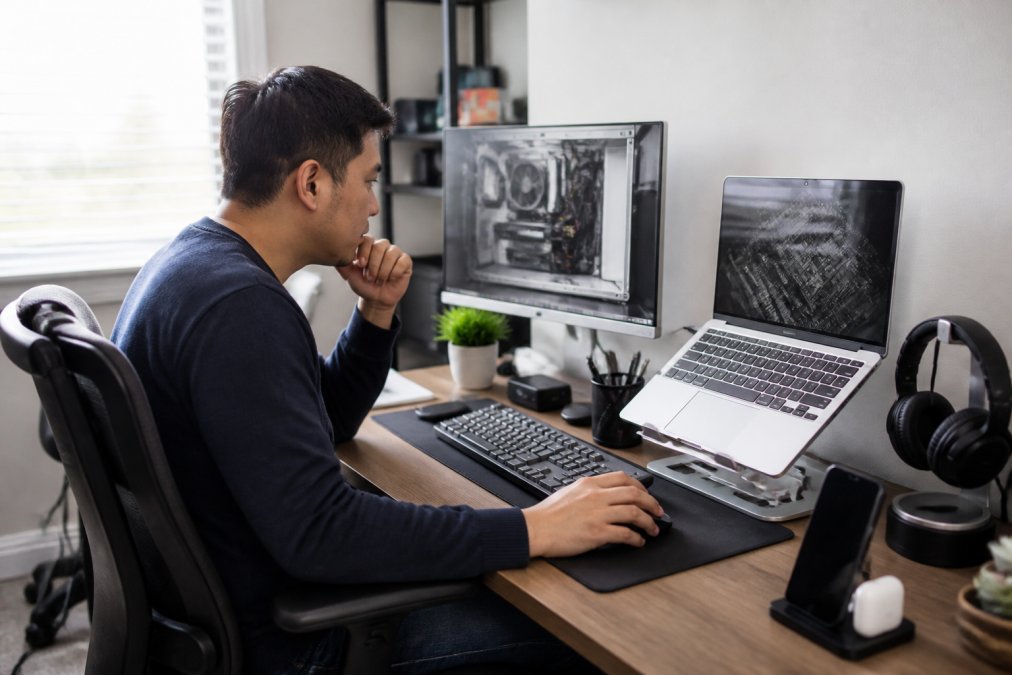 Remote worker at home office desk with multiple monitors, laptop stand, and organized desk accessories, evaluating computer hardware specifications on screen with focused concentration