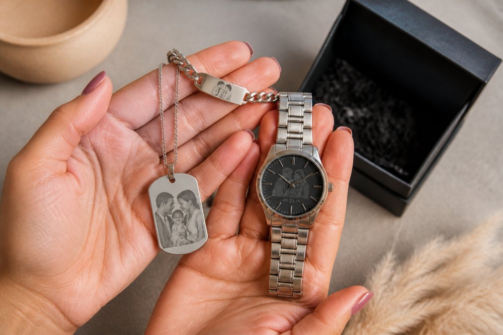 Close-up of hands holding various personalized stainless steel jewelry pieces including an engraved necklace, bracelet, and elegant watch, displayed on a soft neutral background with an open gift box showing branded packaging