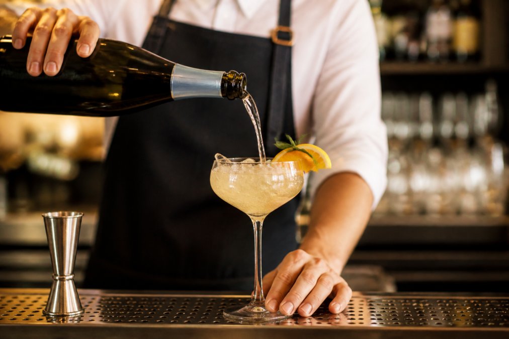Bartender pouring sparkling wine into a coupe glass with fresh citrus garnish and ice, showing the effervescence and bubbles catching light, professional bar setting