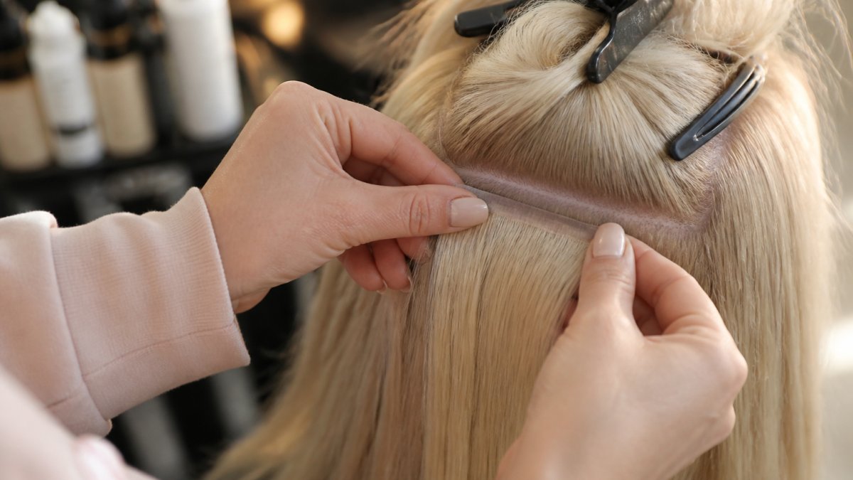 Close-up of a hairstylist's hands lifting a section of fine blonde hair to show a discreet flat weft sitting close to the scalp, soft natural salon lighting