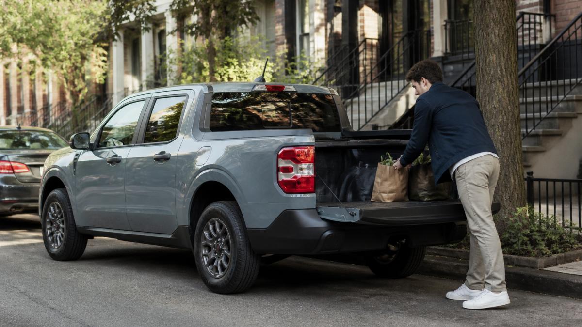 A compact pickup truck parked in a tight urban street parking spot with a driver loading grocery bags into the bed, late afternoon light
