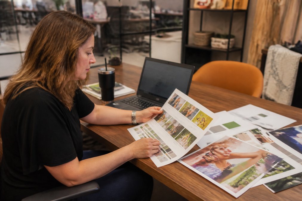 A marketing manager at a retail chain reviewing color proofs of brochures and signage at a desk, with a laptop showing a brand asset management dashboard in the background