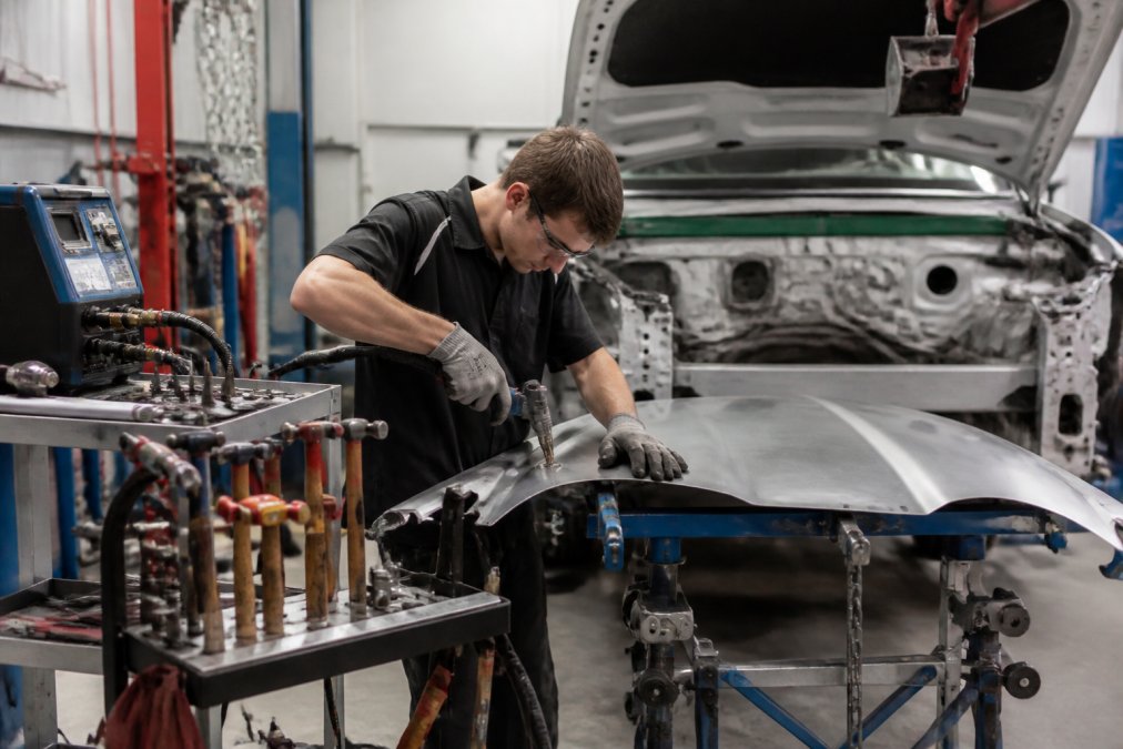 Professional auto body technician working at fully equipped aluminum repair station with stud welding equipment, dolly sets, and specialized hammers visible on organized mobile cart in modern shop environment