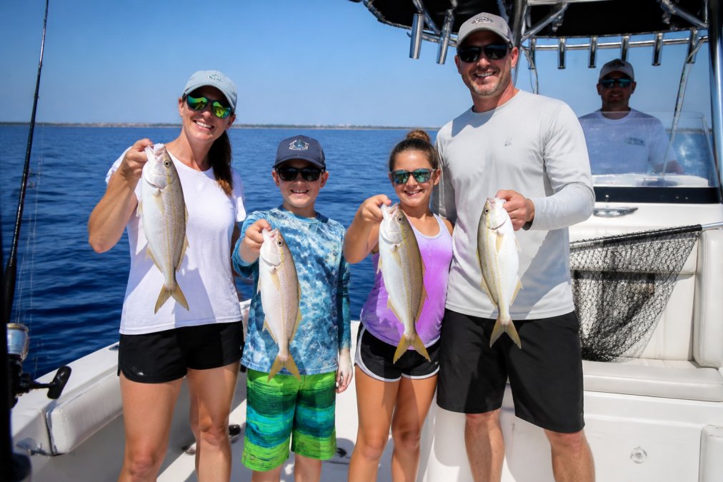 A smiling family of four on a charter boat deck holding up freshly caught fish, bright Gulf Coast blue water and clear skies behind them, captain visible at the helm