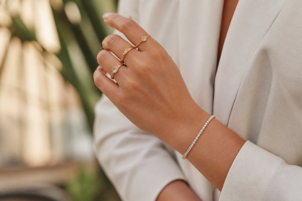 Woman's hand wearing a delicate diamond tennis bracelet and stacked gold rings, photographed in natural daylight with soft focus background