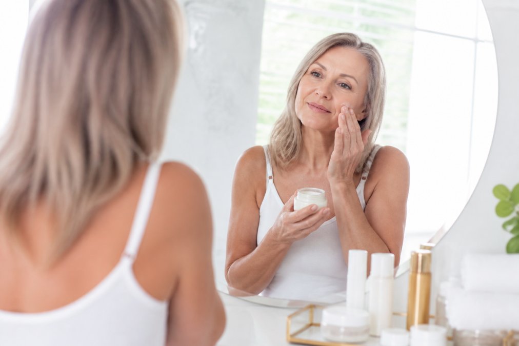 A woman in her fifties applying moisturizer in a bright bathroom, with skincare bottles arranged on the counter, natural light, calm and confident expression