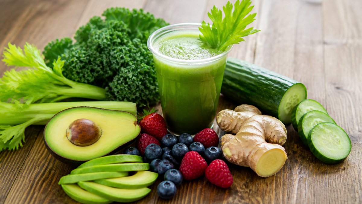 A colorful spread of fresh anti-inflammatory foods on a wooden table, including sliced avocado, berries, cucumber, ginger root, and a glass of green juice