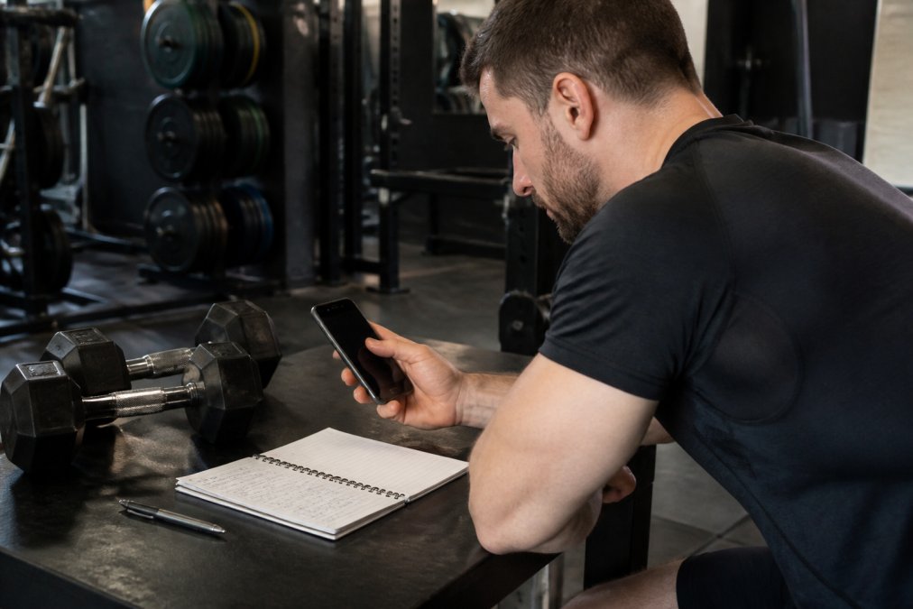 Person at gym workbench reviewing their workout on a smartphone with dumbbells and a training notebook nearby, showing focused attention to tracking details