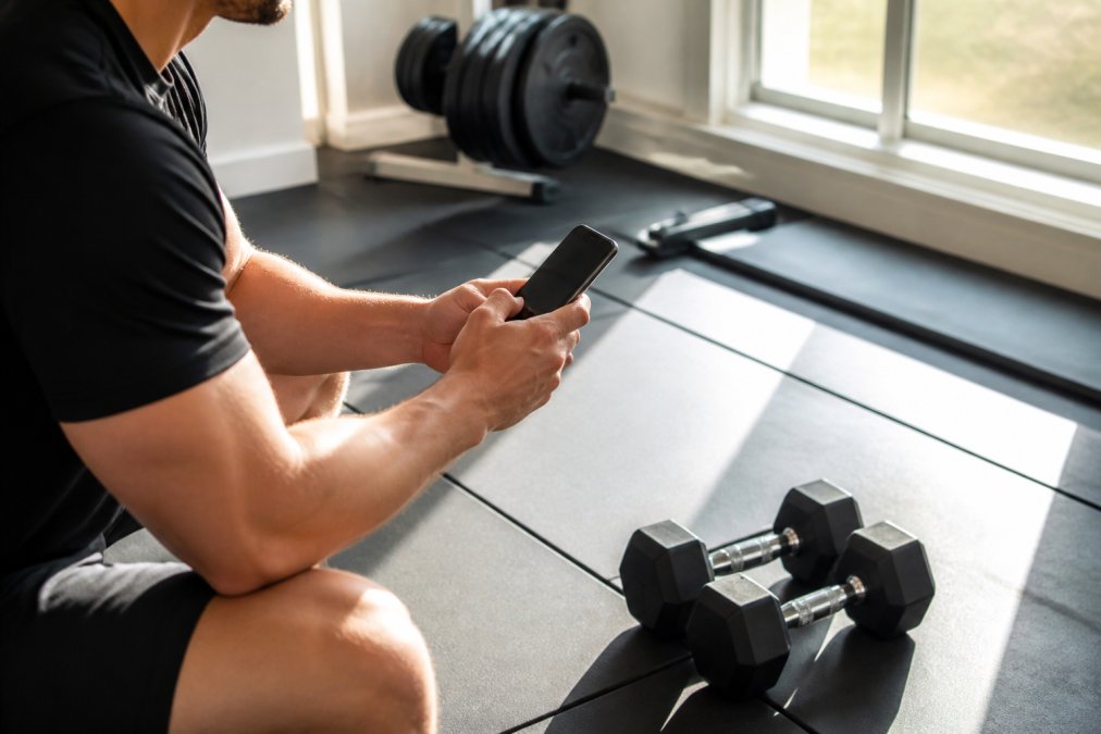 Person holding a smartphone displaying a fitness tracking app while standing next to a set of dumbbells in a bright home gym, warm natural light streaming through a window