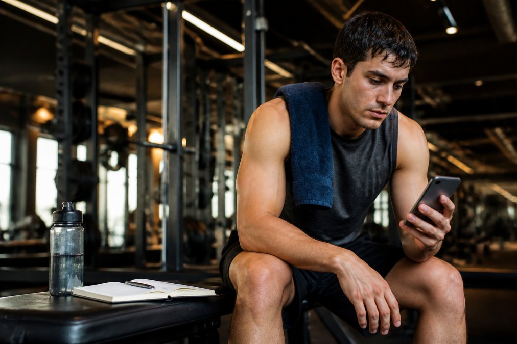 Focused adult checking a workout app on their phone after completing a strength training session, with a notebook and water bottle resting on a gym bench, under warm overhead gym lighting