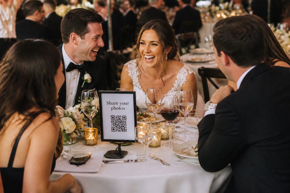 A bride and groom laughing with guests at a reception table, a smartphone on the table displaying a QR code for uploading photos, with a sign that reads 'Share your photos'