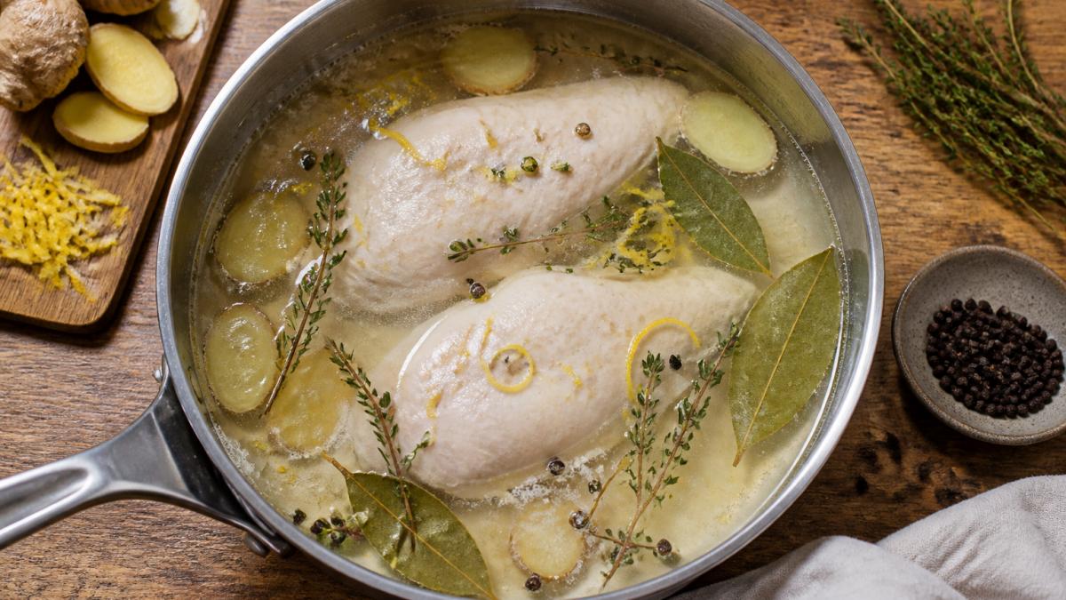 Overhead shot of a shallow pan of gently simmering poaching liquid with chicken breasts, scattered bay leaves, thyme sprigs, sliced ginger, lemon zest and peppercorns on a wooden kitchen counter