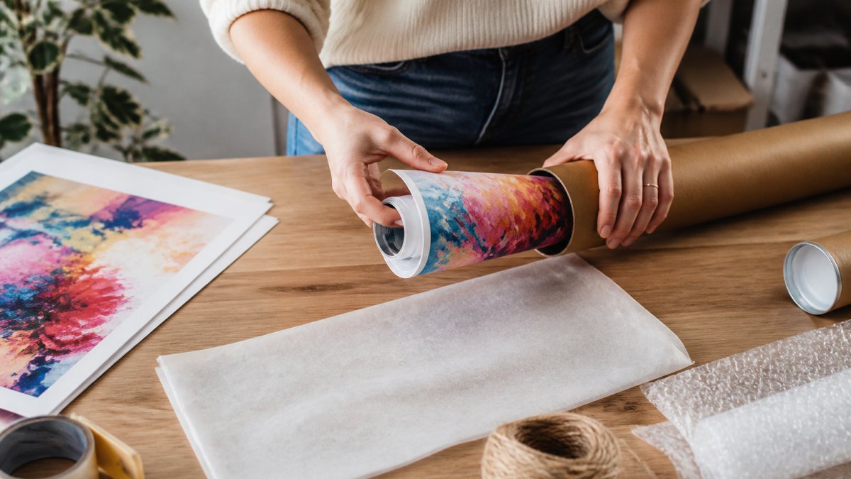 Close-up of hands carefully rolling a colourful art print and placing it into a sturdy cardboard tube on a clean wooden desk, with protective tissue paper nearby