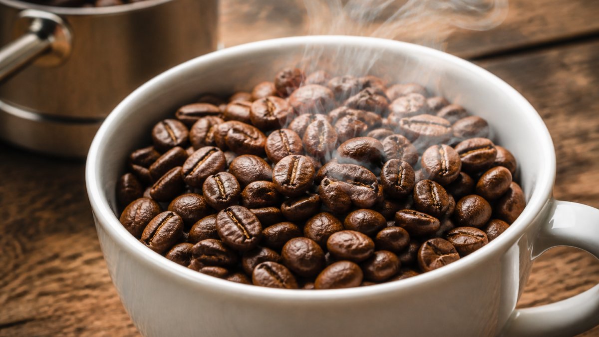 Close-up of freshly roasted medium-roast coffee beans in a white ceramic cup with visible steam rising, showing rich brown color and glossy texture of whole artisan beans