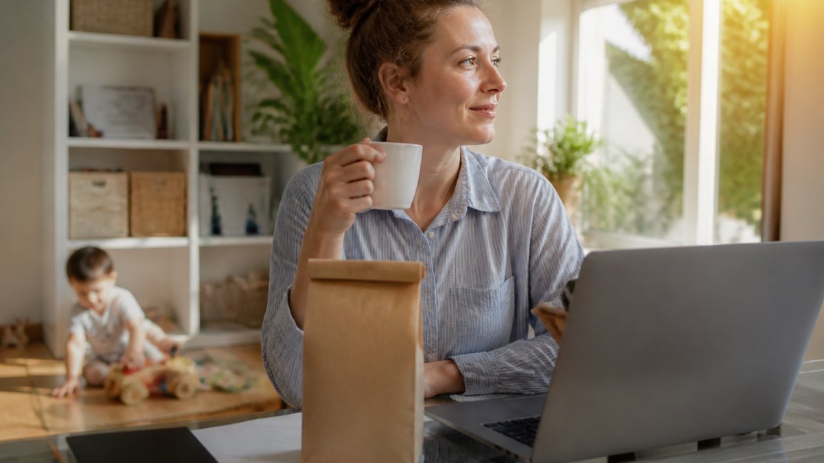Parent in home office holding fresh coffee cup with artisan packaging visible, morning sunlight streaming through window, child's toy visible in background