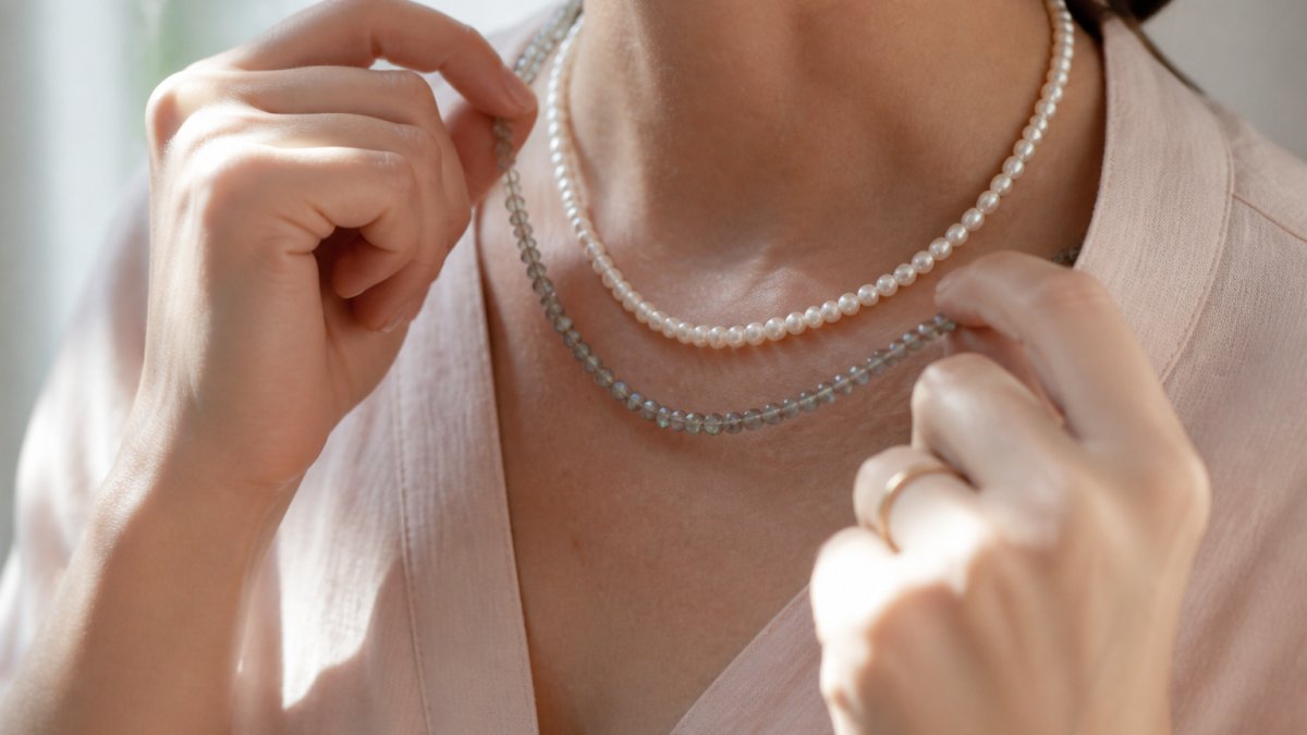 Close-up of a woman's hands layering a dainty labradorite beaded necklace over a light pink pearl necklace, soft natural window light, neutral linen background