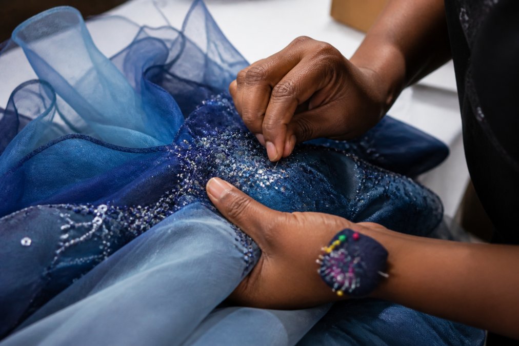 Close-up of artisan hands hand-stitching detailed embellishments and finishing seams on a formal blue ombré organza jumpsuit fabric, showing the precision and care in artisanal production