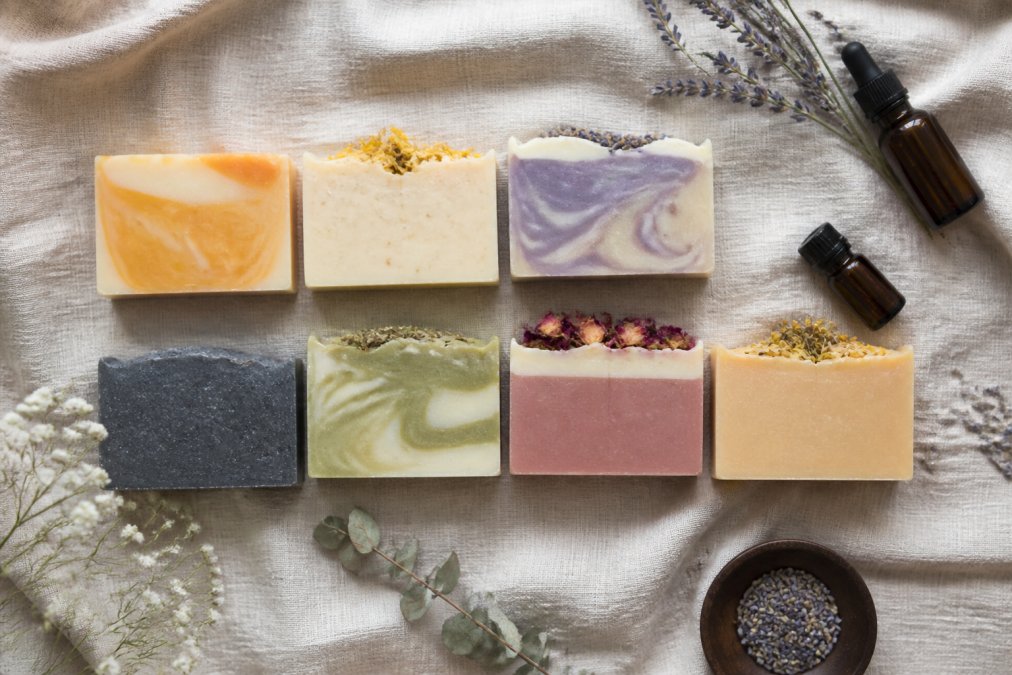 Overhead flat-lay of multiple handcrafted soap bars in various colors and scents arranged on natural linen with dried botanicals, lavender sprigs, and amber essential oil bottles nearby