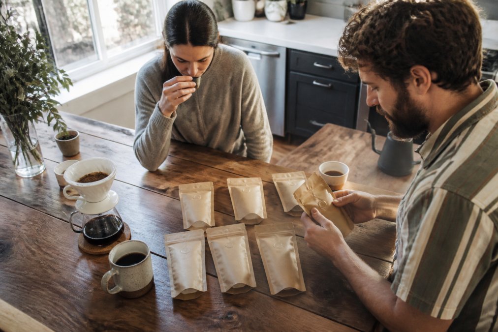 Overhead shot of six small 2oz coffee sample bags arranged on a rustic wooden kitchen counter beside a ceramic pour-over dripper and a steaming mug of black coffee, soft morning window light