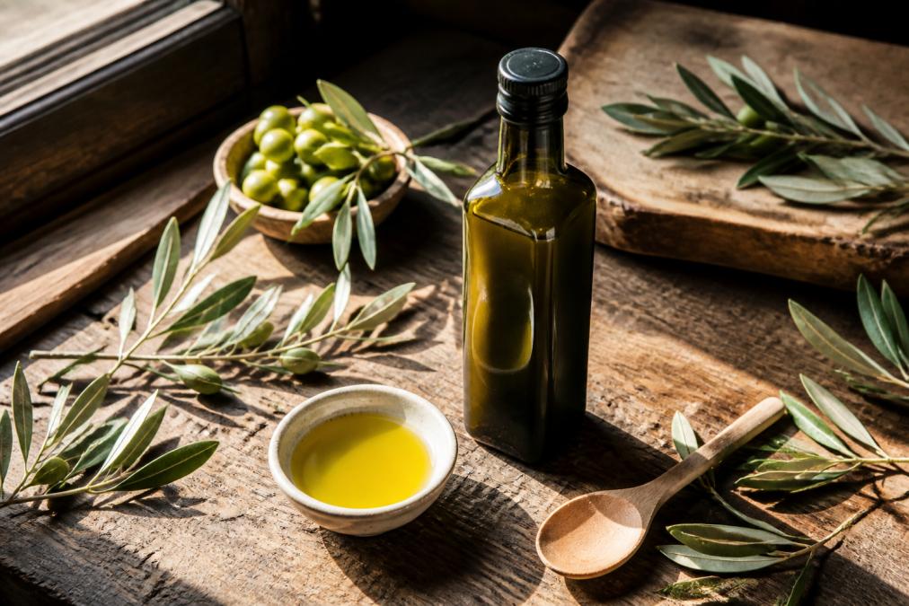 Overhead shot of a rustic kitchen bench with a dark glass bottle of cloudy green olive oil beside a small bowl of freshly pressed oil, olive branches and a wooden spoon, warm afternoon light through a window