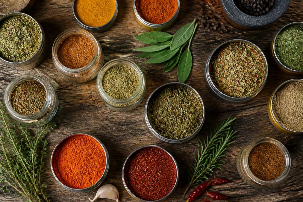 Overhead flat lay of open artisanal spice jars and tins with colorful spice blends, herbs, and hand-blended seasonings arranged on a rustic wooden surface with warm natural lighting