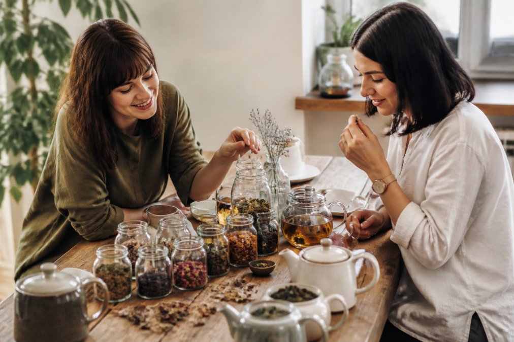 Overhead shot of artisanal loose leaf tea collection displayed in glass jars with various colored teas, fresh botanicals, and handwritten flavor labels on a wooden surface