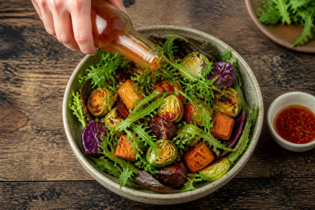 A vibrant salad bowl with mixed greens, roasted vegetables, and a glossy chilli vinegar dressing being drizzled over the top, shot from above on a rustic table
