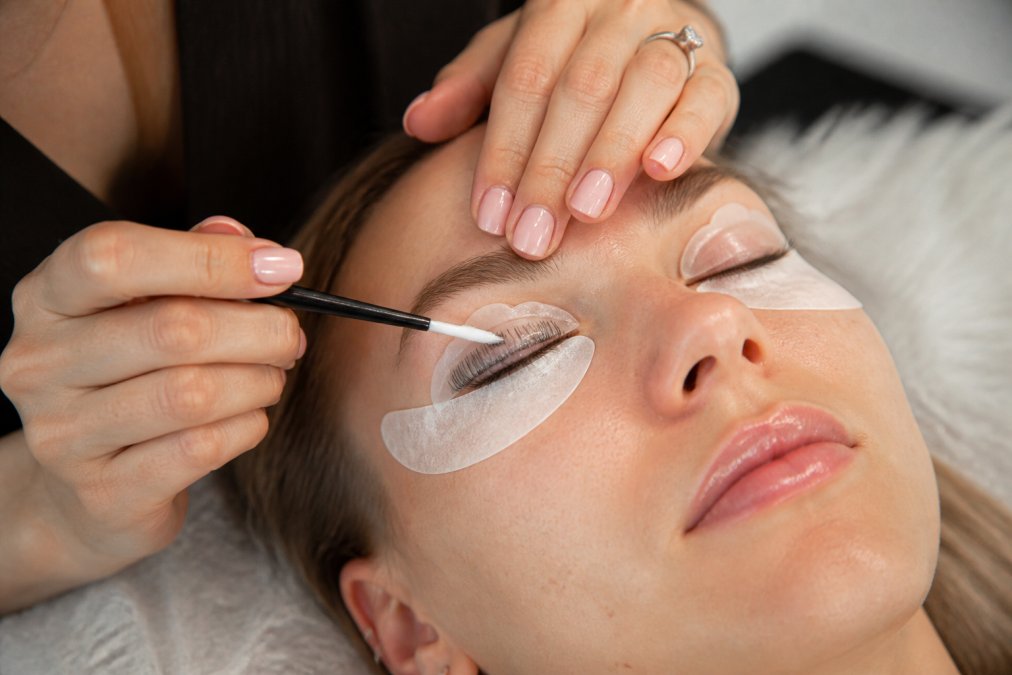 Close-up of hands carefully applying lash lift solution to natural eyelashes using an applicator brush, with silicone shields properly positioned on the lash line