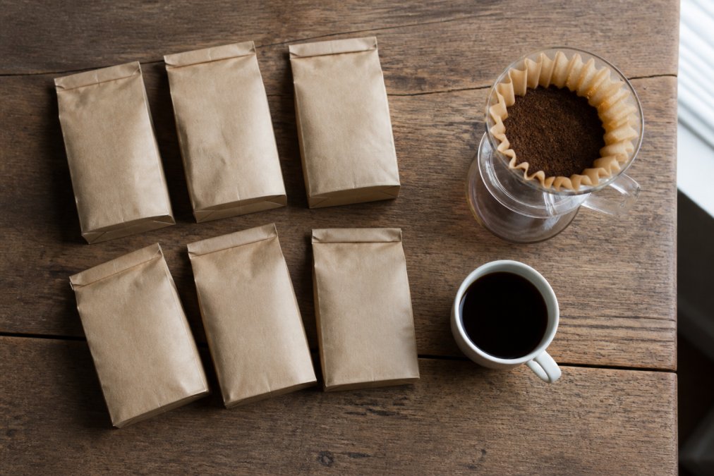 Six coffee sample bags arranged next to a pour-over dripper and a cup of black coffee on a wooden table.