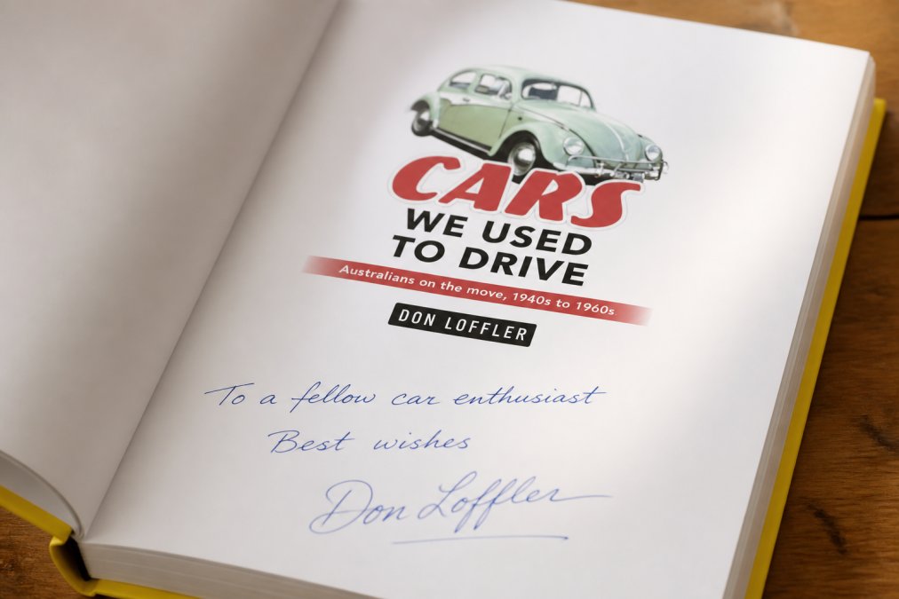 Close-up of an open automotive book showing author's handwritten signature and personal inscription on the title page, with warm natural lighting illuminating the page