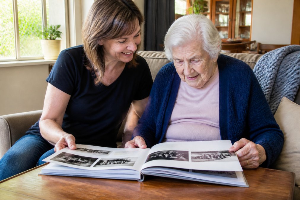 Carer or family member sitting beside an elderly person, both looking at a decade-specific photo book together, sharing a moment of connection and genuine conversation