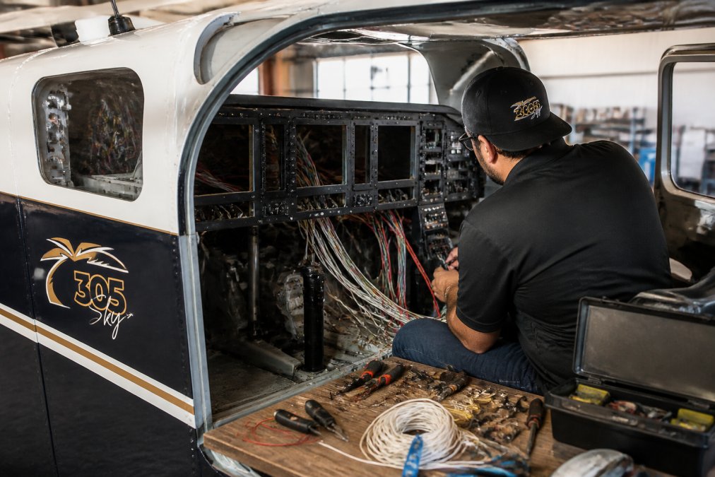 Aircraft technician in maintenance hangar working on panel-mounted avionics equipment with multiple screens, wiring harnesses, and specialized tools visible on workbench under bright overhead lighting