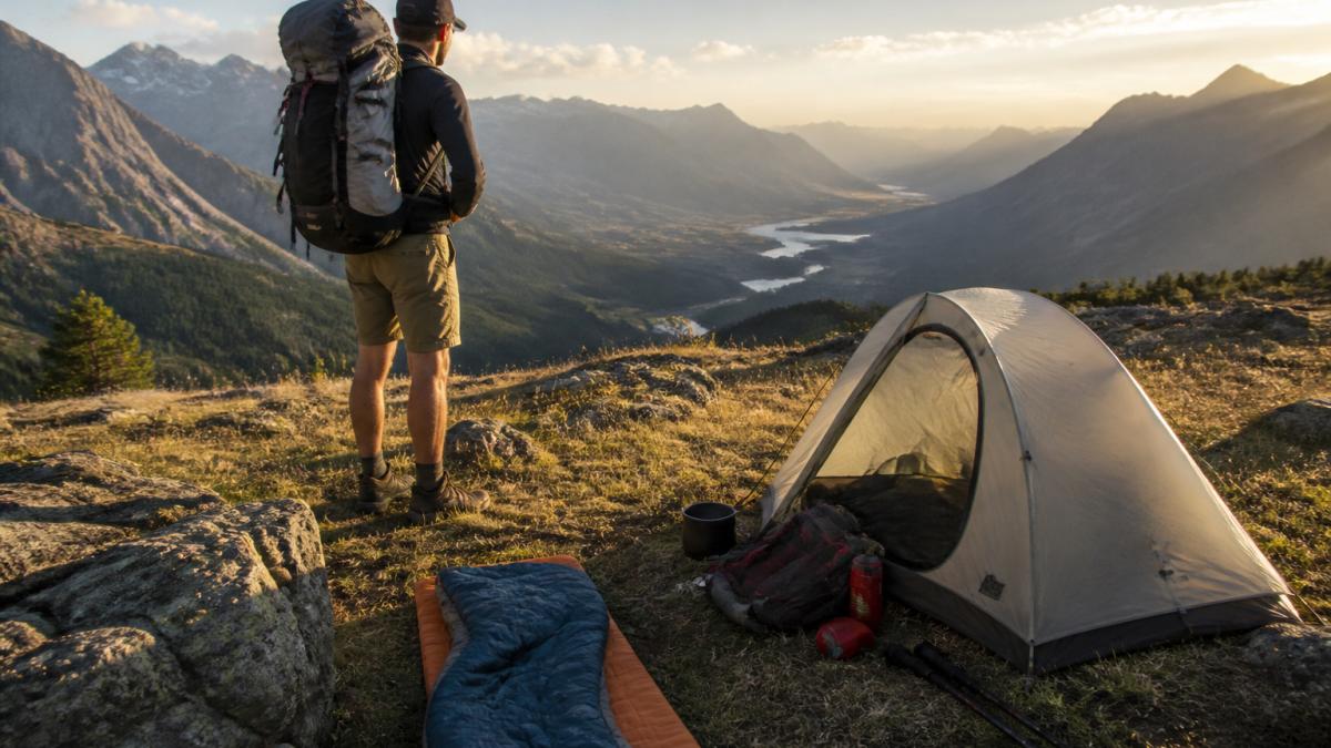 Backpacker with loaded hiking backpack standing on a mountain trail overlooking a valley, tent and sleeping gear visible on the ground in morning light