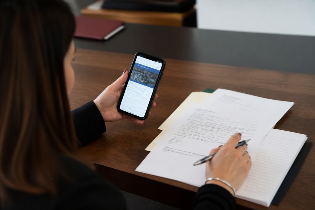 Close-up of hands holding phone showing legitimate bail bond company website and official documentation, person reviewing paperwork on desk with pen