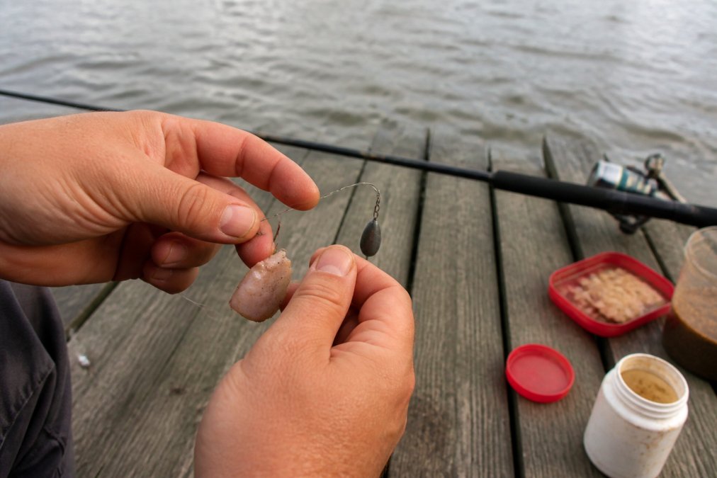 Close-up of hands tying a fishing rig with sinker, hook, and bait on a wooden dock with river water visible in the background