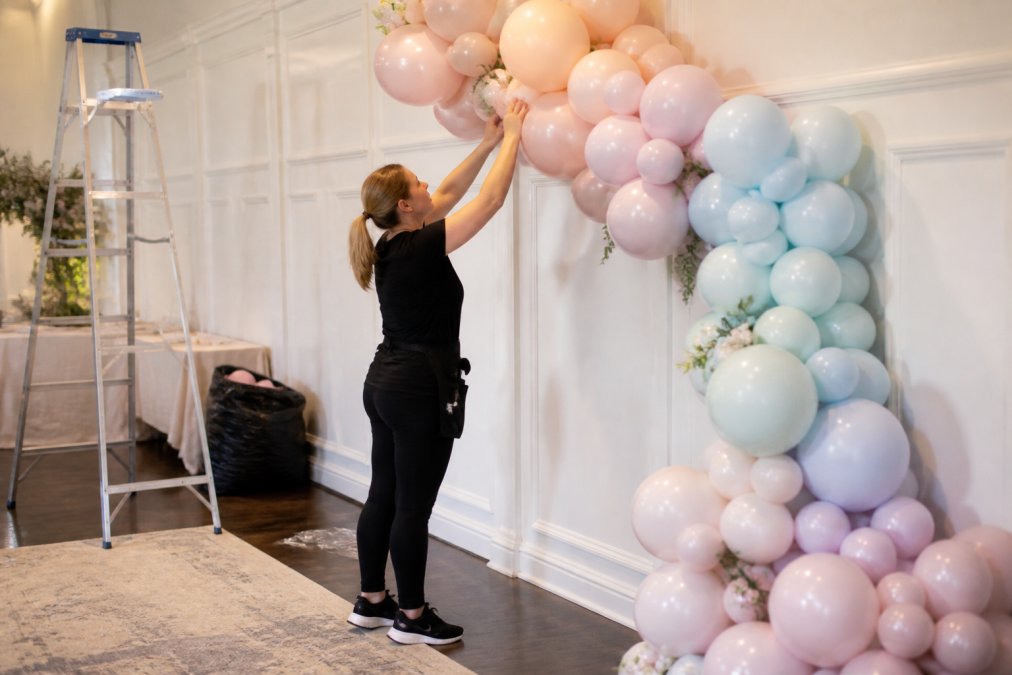A professional balloon decorator arranging a large organic balloon garland in pastel colors against a white venue wall, with ladders and supply bags visible in the background