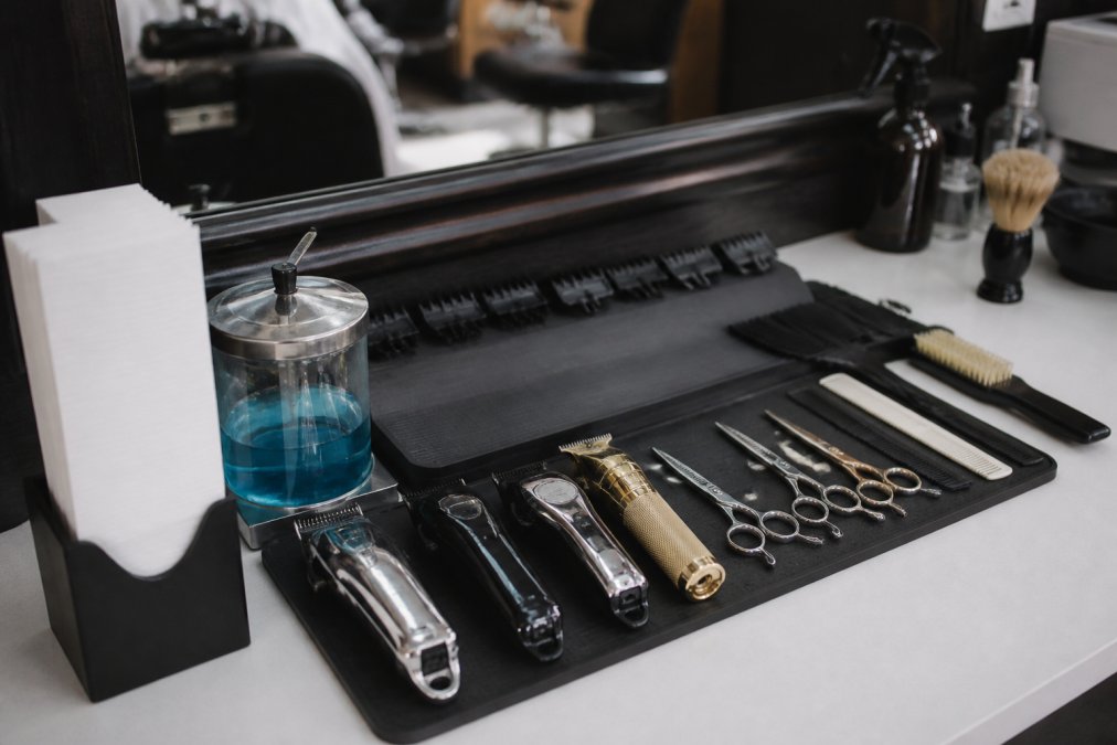 Professional barber station with clippers, scissors, and tools neatly arranged on a magnetic mat with neck strips in a holder and a disinfecting jar visible, showing a well-organized workspace ready for client service