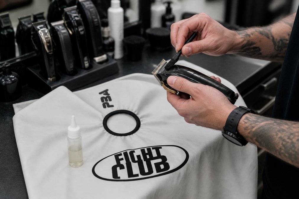 Barber hands demonstrating blade cleaning and maintenance with clipper oil and soft brush at a professional station, showing proper sanitization technique with natural lighting