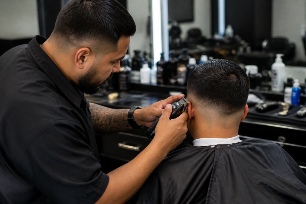 Professional barber performing a precise fade on a client using a cordless clipper, showing proper ergonomic grip and technique at a barbershop station with organized tools visible in the background