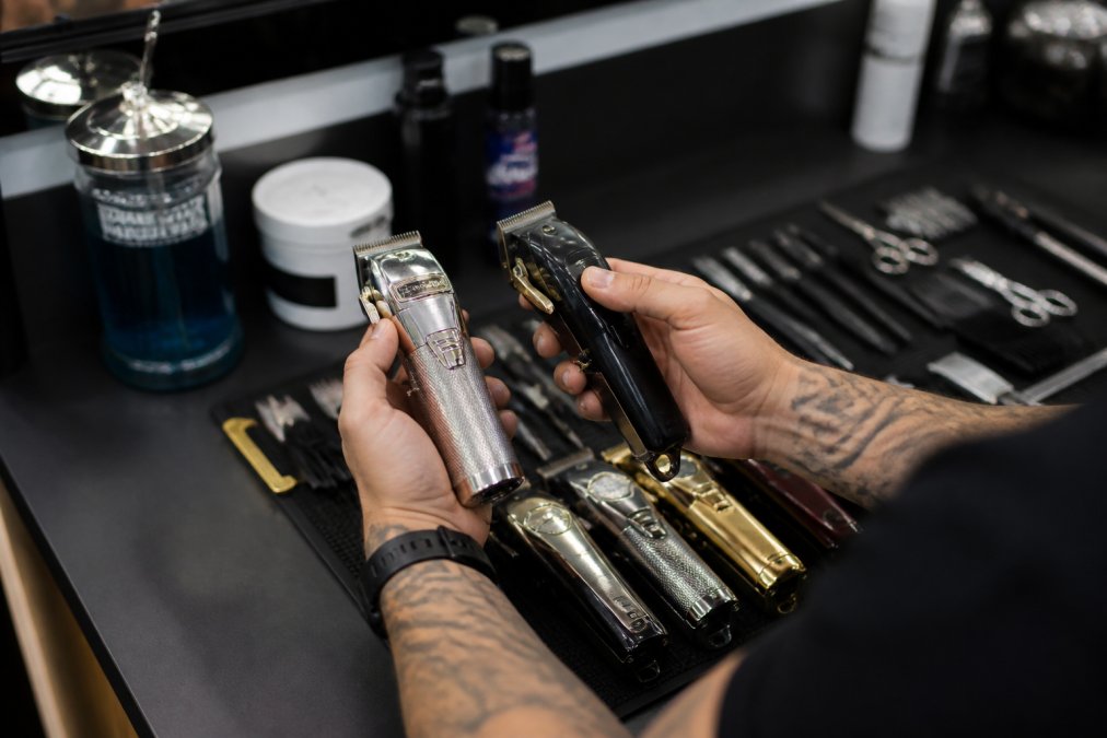 Professional barber's hands holding precision clippers and trimmers over an organized barber station with disinfecting jar and clean tools