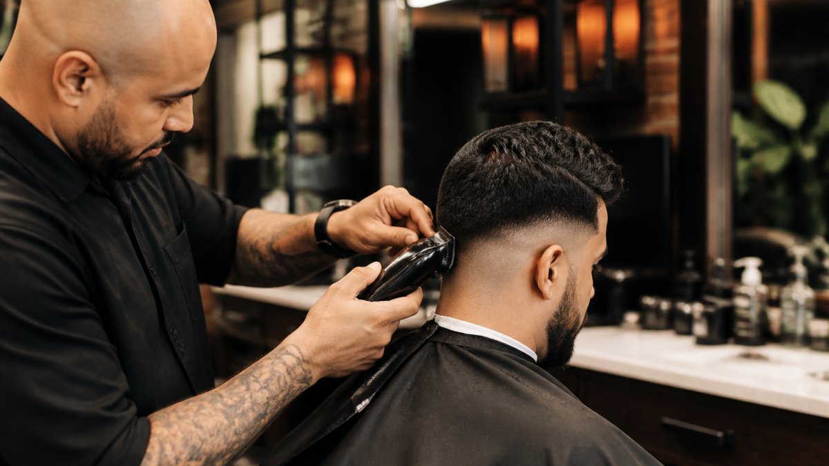 Professional barber performing precision fade haircut on client, using clippers with focused technique and attention to detail in modern barbershop setting with warm lighting