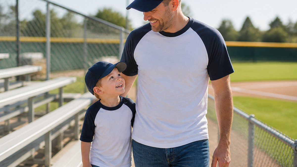 Toddler boy and his father wearing matching baseball shirts while smiling at a sunny stadium game day
