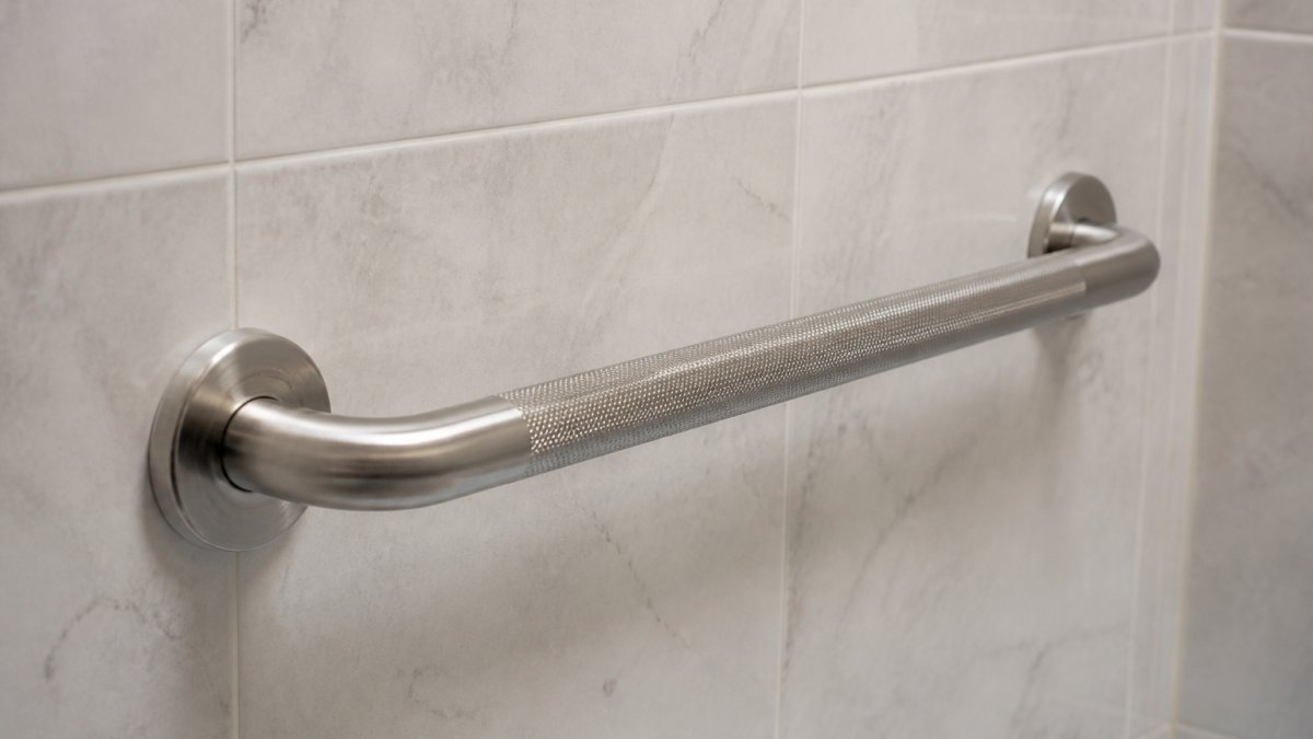 A close-up photograph of a stainless steel grab bar mounted on a clean, tiled shower wall in a well-lit bathroom, showing the secure mounting and textured grip surface