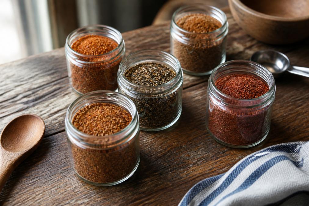 A rustic wooden table holding several open 5 oz jars of BBQ rub with visible spice textures in brown, red, and black tones, natural window light